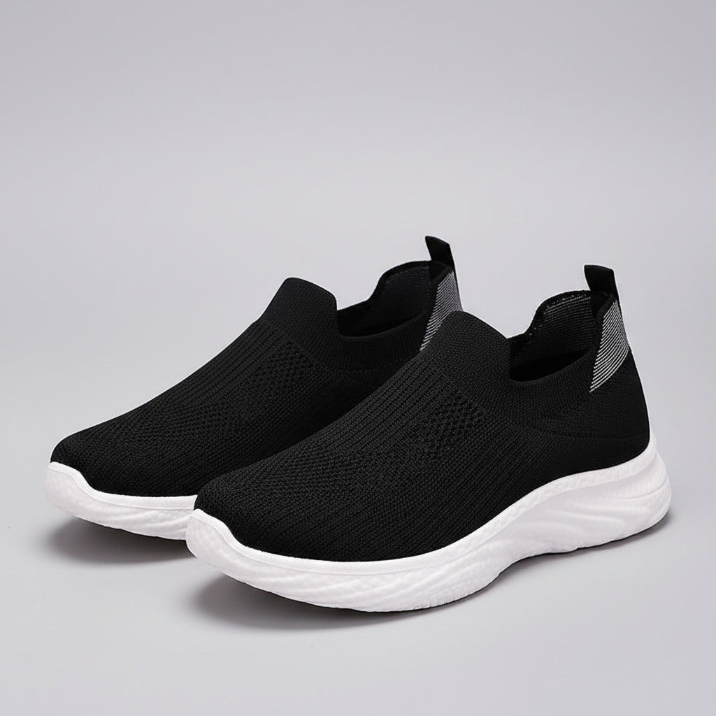 Black slip-on sneakers with white soles on a wooden floor.